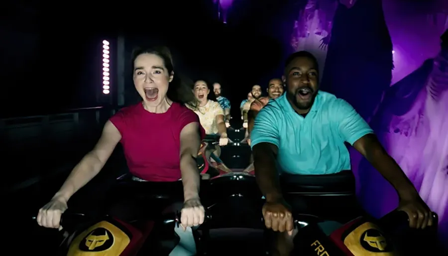A group of excited and screaming riders are experiencing a thrilling moment on an indoor roller coaster, with expressions of exhilaration and enjoyment under dim lighting.