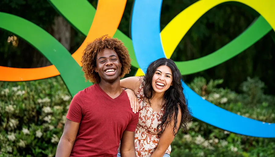 Two people are smiling and posing in front of a colorful, abstract sculpture.