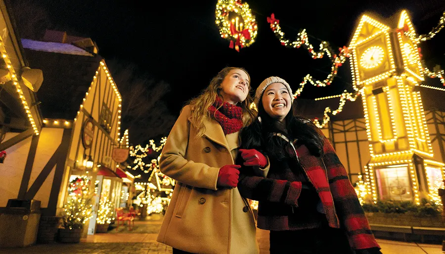 Two people are smiling and looking upwards while enjoying a festive outdoor setting adorned with Christmas lights and decorations at night.