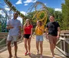 A group of four friends are walking and laughing together on a wooden bridge with a roller coaster in the background on a sunny day