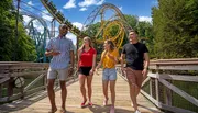 A group of four friends are walking and laughing together on a wooden bridge with a roller coaster in the background on a sunny day.
