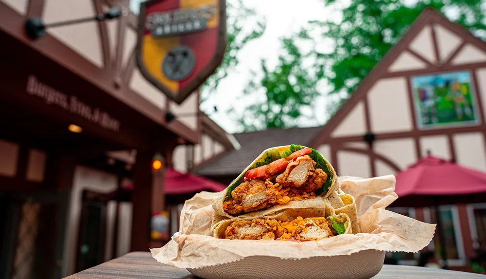 A close-up of a sliced wrap filled with fried chicken lettuce and tomatoes sits on a table outside a medieval-style building