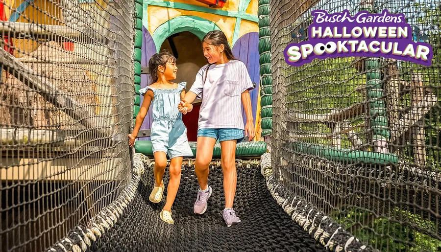 Two young girls are happily walking hand in hand on a netted bridge at Busch Gardens' Halloween Spooktacular.