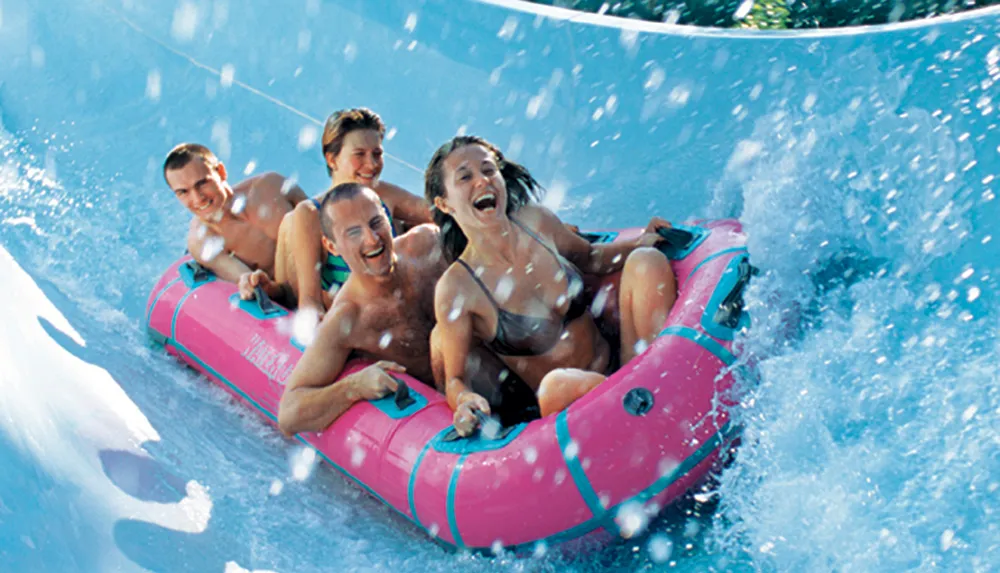 Four people are enjoying a thrilling ride down a water slide in a pink inflatable raft splashing water around and laughing
