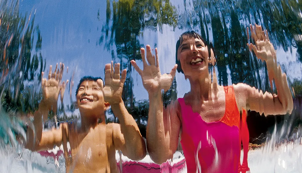 A young boy and a woman are smiling and reaching out towards the camera through a clear curtain of water likely enjoying a sunny day at a water play area