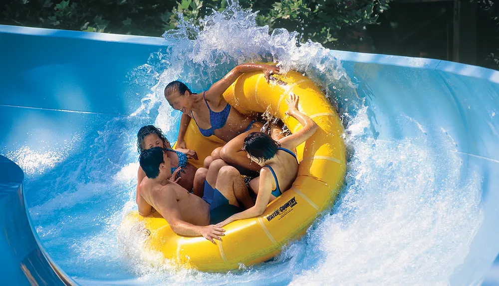 Several people are joyfully riding down a water slide in a large yellow raft surrounded by splashing water