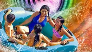 A group of people, including children, are enjoying a ride on a vibrant, colorful water slide in a blue raft, surrounded by sprays of water.