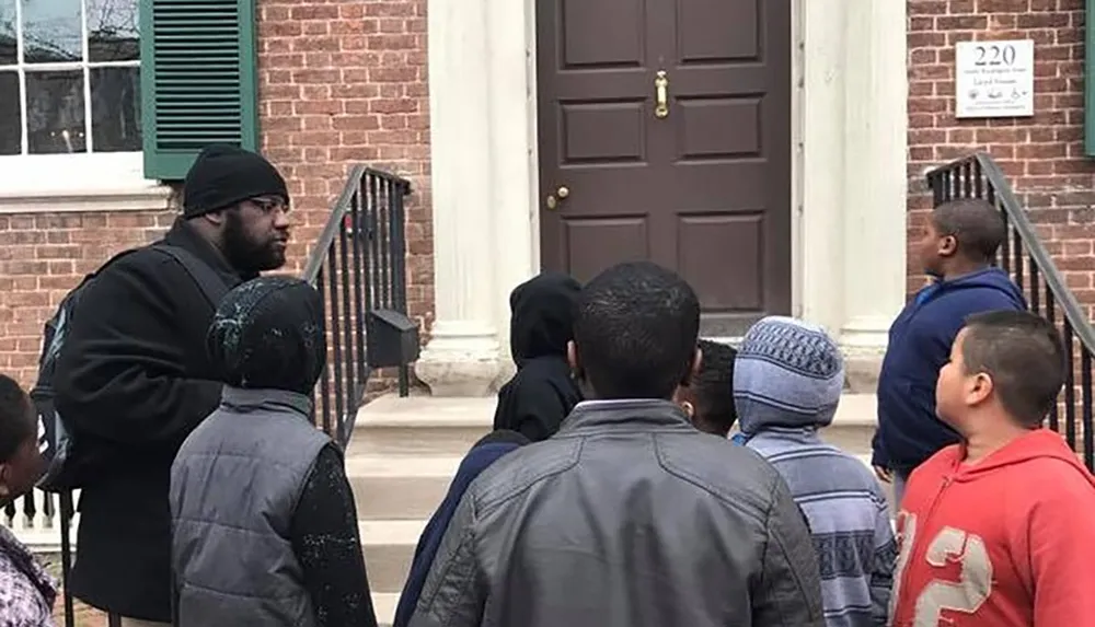 A group of children listens to an adult speaking to them on the steps of a brick building with a brown door marked with the number 220