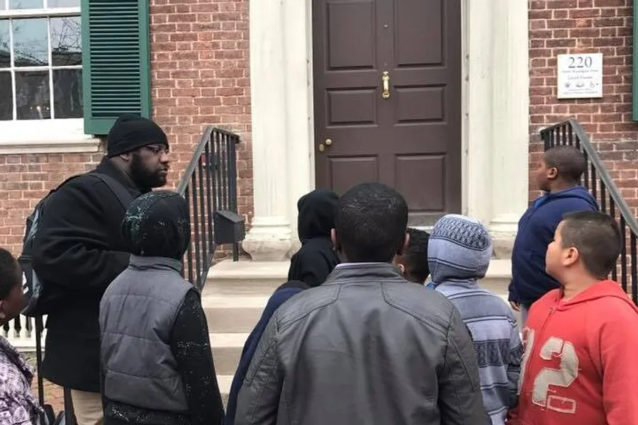 A group of people is gathered outside a building, listening to a man who appears to be speaking or guiding them.