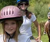 A young girl in the foreground with a pink helmet smiles during a horseback riding session with two other riders in the background