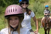 A young girl in the foreground with a pink helmet smiles during a horseback riding session, with two other riders in the background.