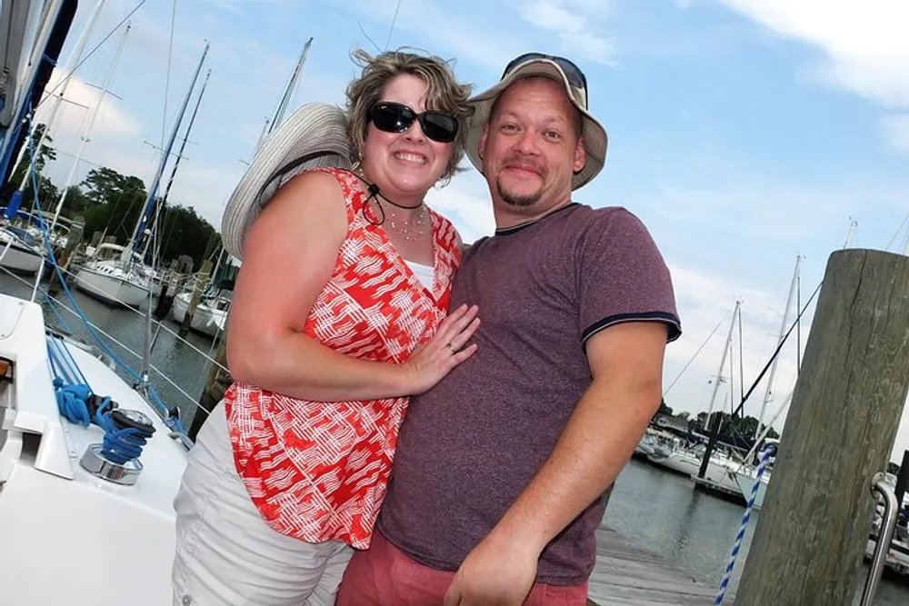 A smiling couple is posing together at a marina with sailboats in the background