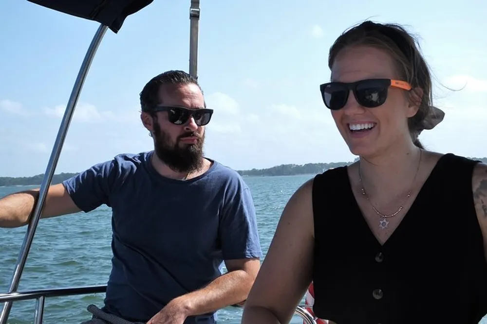 A man and a woman wearing sunglasses are enjoying their time on a boat with the man steering and the woman smiling brightly