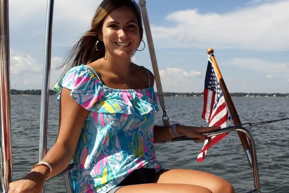 A smiling woman in a colorful top is sitting on a boat with a small American flag in the background