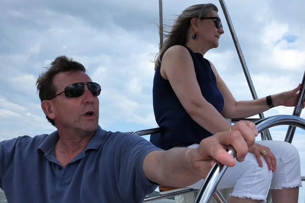 A man and a woman both wearing sunglasses are on a boat the man is steering with a concerned expression while the woman sits relaxed in the background