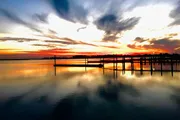 The image displays a serene sunset over a calm body of water with a silhouette of a pier and clouds reflected on the water's surface.