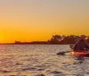 A person in a yellow kayak is paddling near the shore at sunset while a horse and its rider are visible in the background creating a serene silhouette against the glowing sky