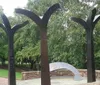 The image shows a group of abstract tree-like metal sculptures in a park-like setting with lush greenery in the background