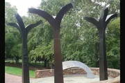 The image shows a group of abstract, tree-like metal sculptures in a park-like setting with lush greenery in the background.