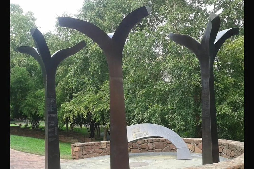 The image shows a group of abstract tree-like metal sculptures in a park-like setting with lush greenery in the background