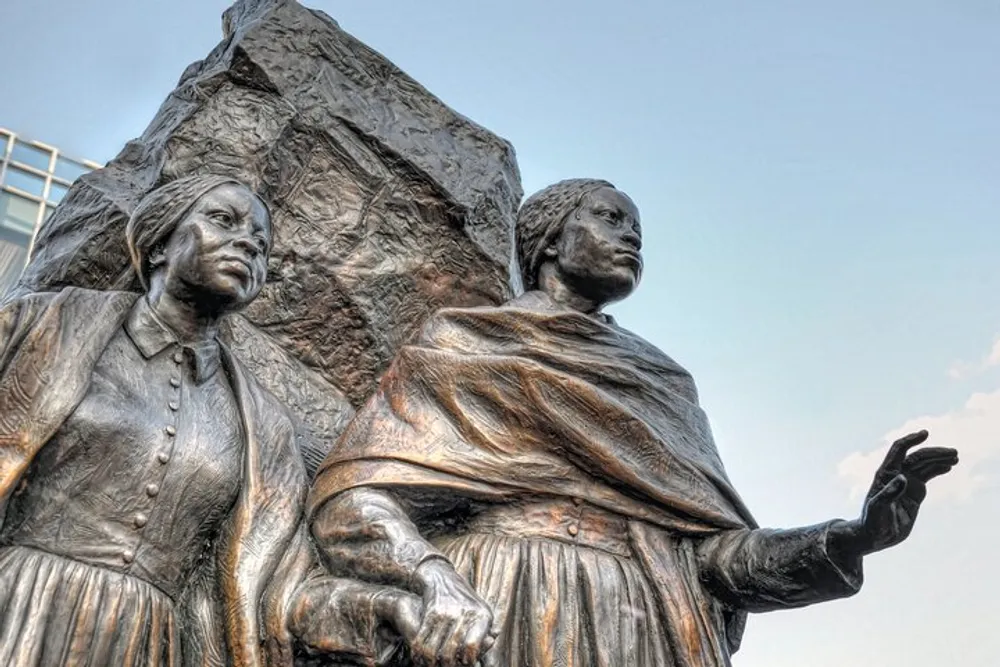 The image shows a bronze statue of two women one standing with an outstretched hand and the other seated against a backdrop of a clear sky