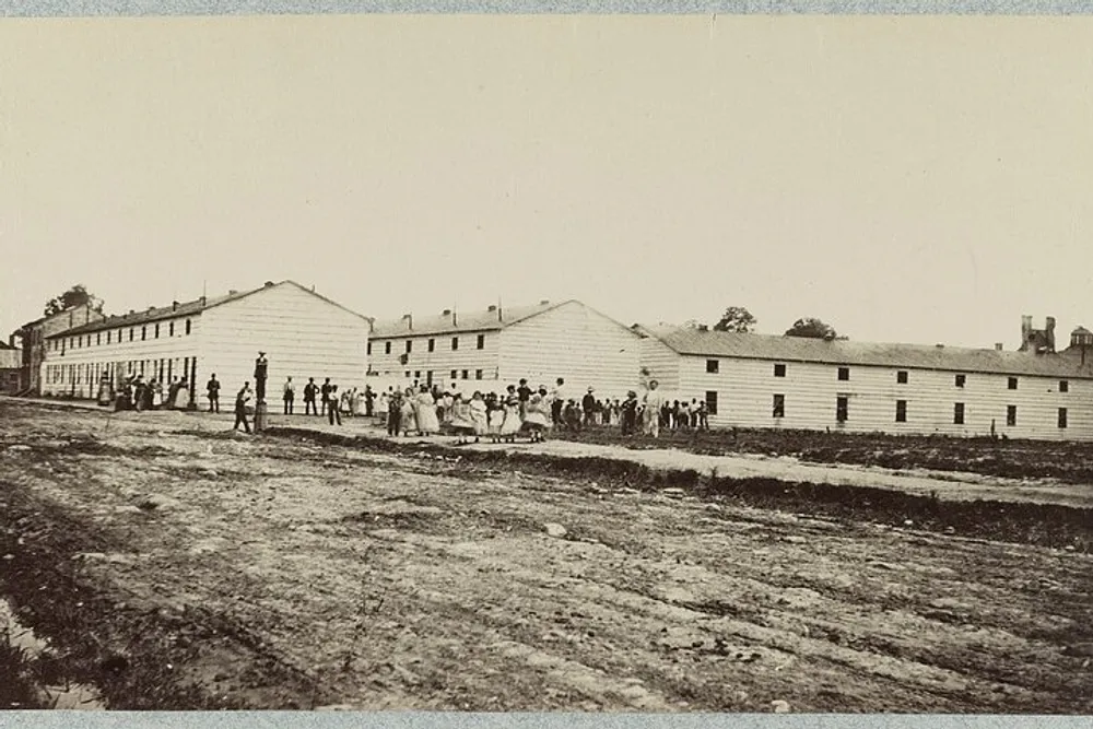 The image shows a historical black-and-white photograph of a group of people standing near long barrack-like buildings with a dirt road in the foreground