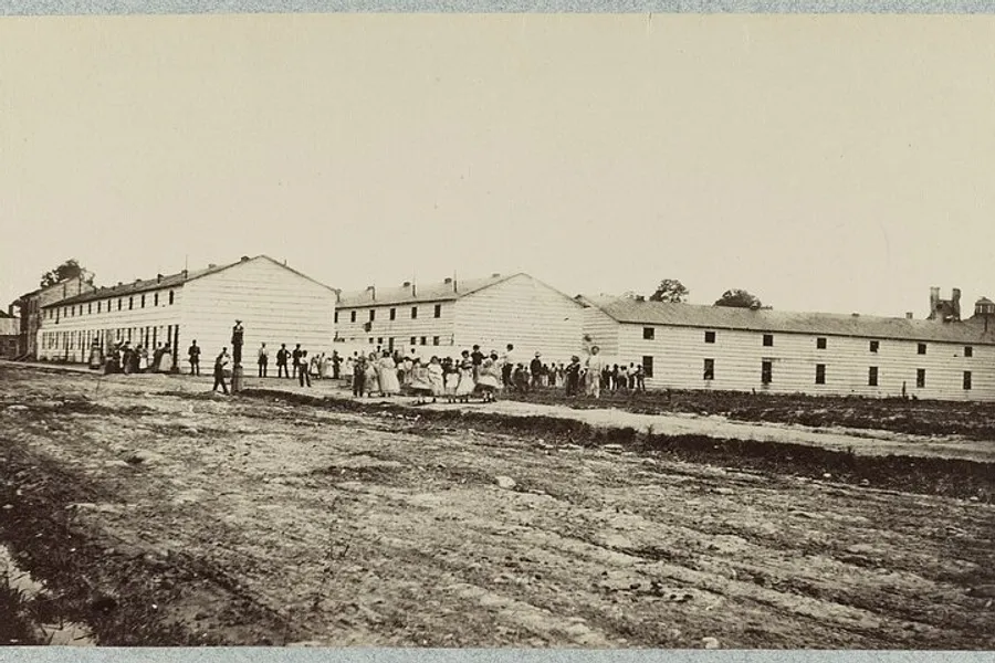 The image shows a historical black-and-white photograph of a group of people standing near long barrack-like buildings with a dirt road in the foreground.
