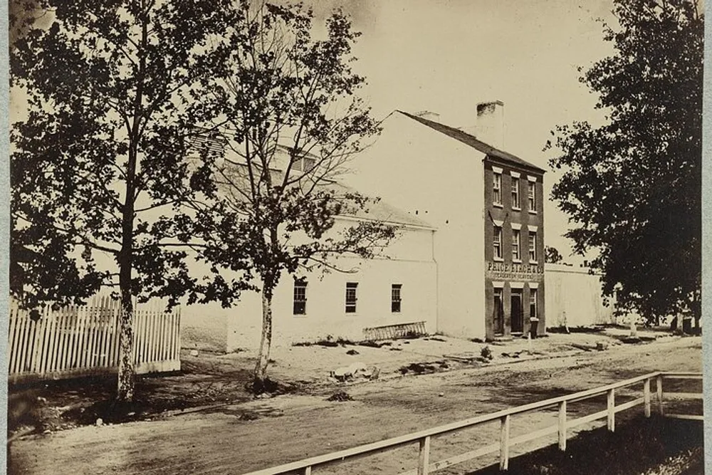 The image shows a vintage black and white photograph of a three-story building with PHOTOGRAPHIC written on its side adjacent to a white single-story structure both behind a white picket fence and surrounded by trees