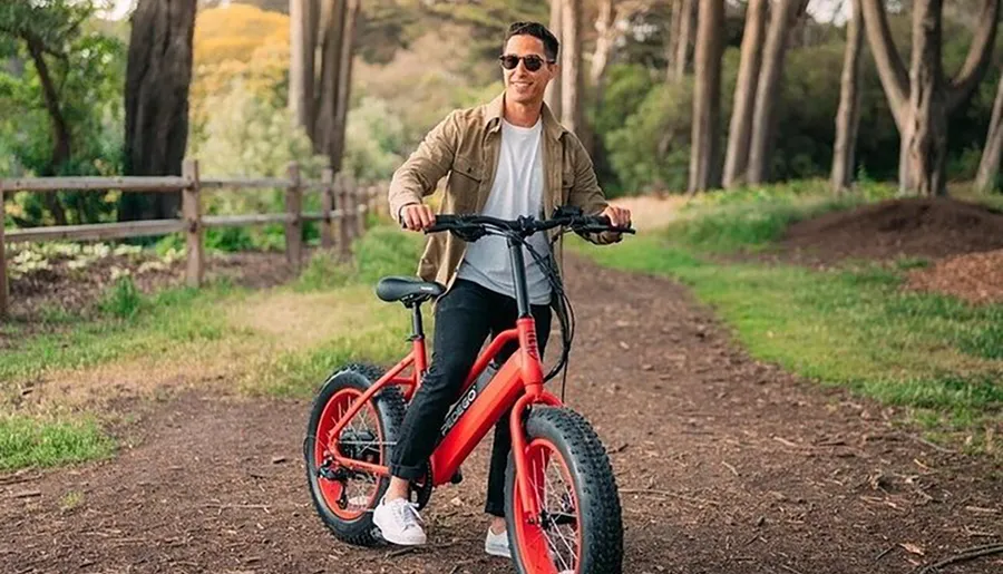 A person is smiling while standing over a red electric bicycle on a dirt path in a wooded area.