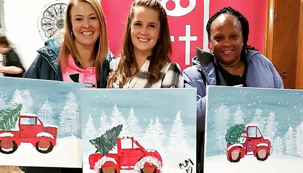 Three people are smiling and holding up their paintings of a red pickup truck with a Christmas tree in a snowy landscape