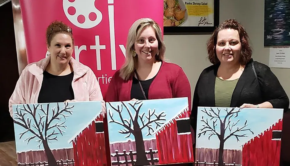 Three people are proudly displaying their similar paintings of a tree against a blue and red background at what appears to be a painting class