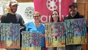 Four individuals are standing side by side, each proudly holding up a colorful painting of a tree scene.