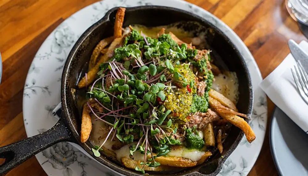 The image shows a skillet of poutine a dish consisting of fries and cheese curds topped with meat and green garnish served on a floral-patterned plate on a wooden table