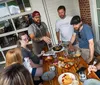 A group of people is engaged in outdoor dining with one person explaining or serving a dish at the table