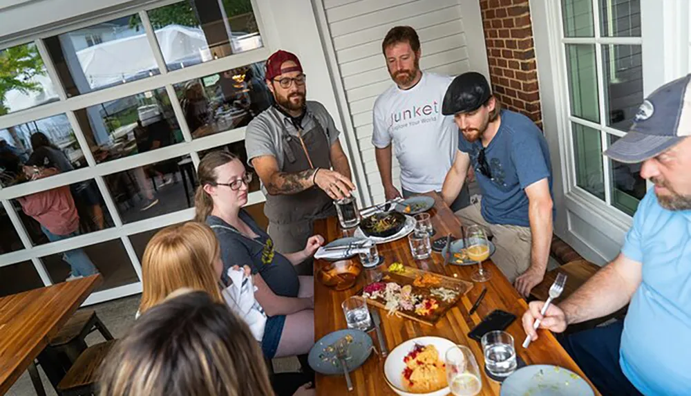 A group of people is engaged in outdoor dining with one person explaining or serving a dish at the table