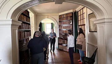 A group of people is on a tour in an elegant room with arched doorways and wall-to-ceiling bookshelves filled with old books.