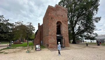 Visitors explore the ruins of a historic brick building with an adjacent large tree, set against a cloudy sky.