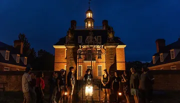 A group of people stand in front of an illuminated historic building at dusk, listening to a guide dressed in period clothing.