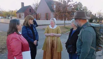 A group of people is listening to a woman dressed in historical attire, possibly a reenactor or tour guide, on a street that appears to come from an earlier era.