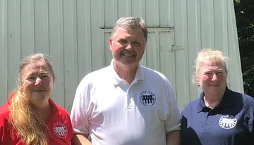 Three people are smiling and posing for a photograph in front of a white corrugated metal backdrop, with two of them wearing shirts with similar circular logos.