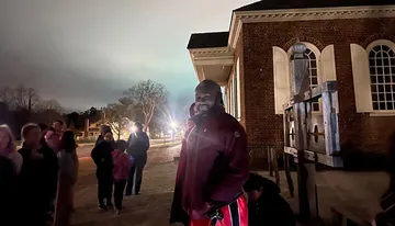 A person is smiling for the camera during a night-time outdoor event near an historic-looking building, with other people and a wooden stock in the background.