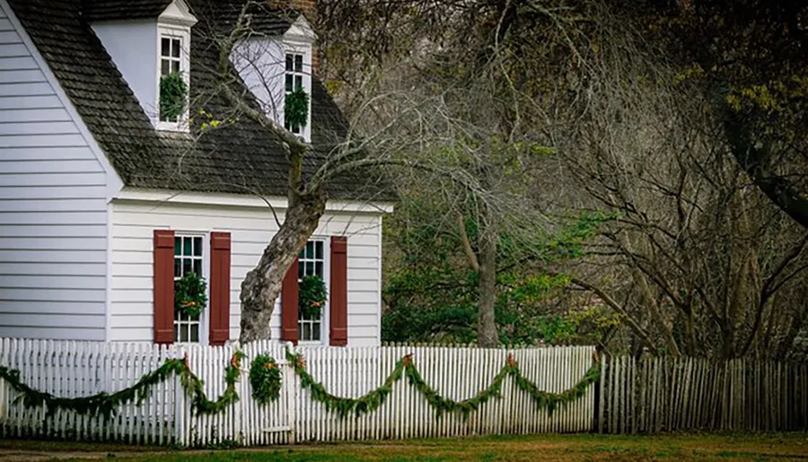 This image features a traditional white house with red shutters behind a white picket fence adorned with greenery, set against a backdrop of leafless trees.