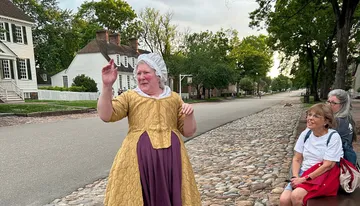 A person dressed in historical costume is gesturing while speaking to onlookers on a cobblestone street lined with colonial-style houses.