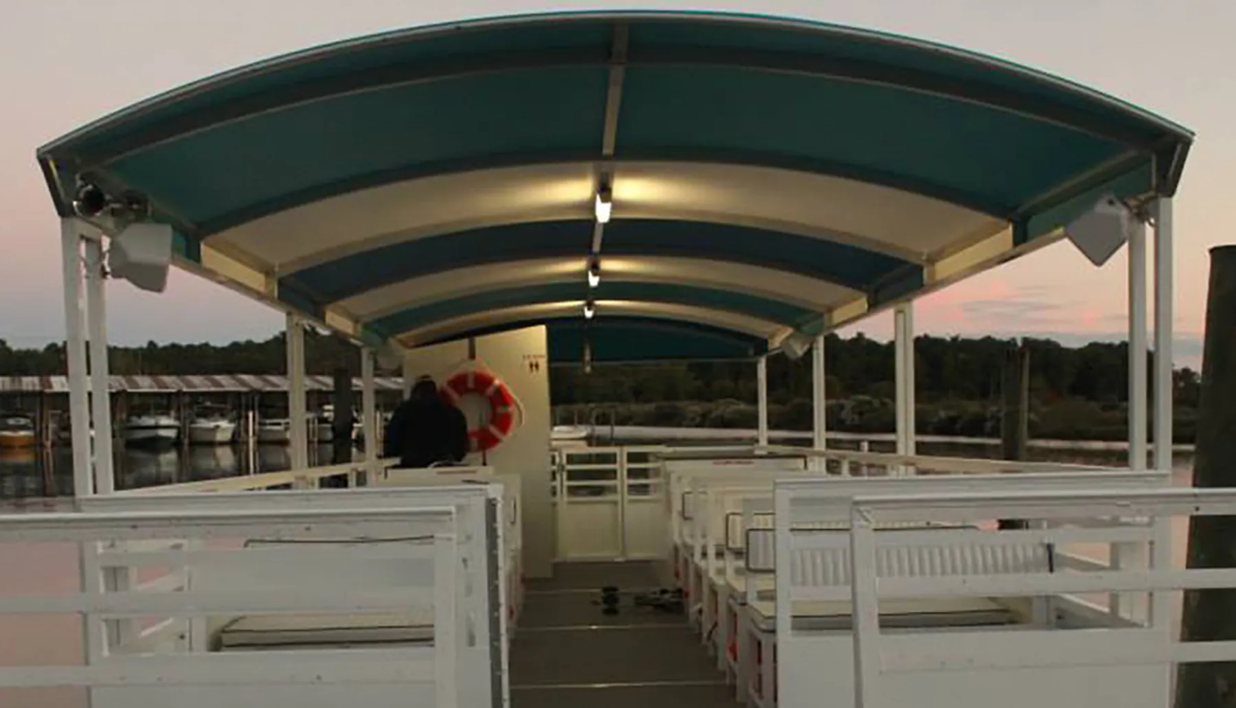 The image shows the inside of a covered, open-air boat with rows of white benches, docked near a marina at sunset.