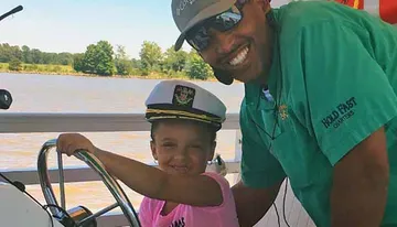A man and a child, both smiling, are steering a boat with the child wearing a captain's hat.