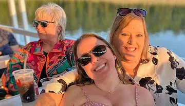 Three women are smiling and enjoying a sunny day on a boat with drinks.