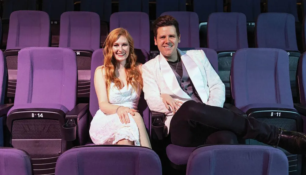 A smiling woman and man are sitting casually in the purple seats of an empty theater