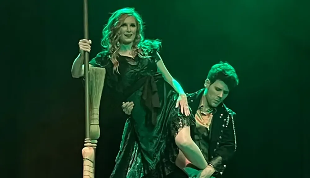 A woman in a black dress stands holding a large broom while being lifted by a man in a studded jacket on a stage with green lighting