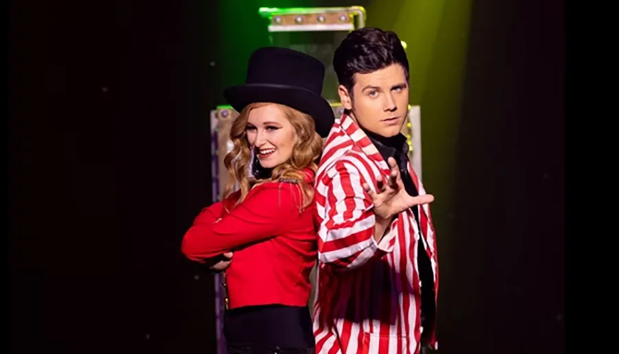 A woman in a red jacket and top hat stands back-to-back with a man in a red and white striped jacket, gesturing dramatically.