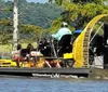 Three people wearing headphones are seated in a boat gliding on a river under a partly cloudy sky